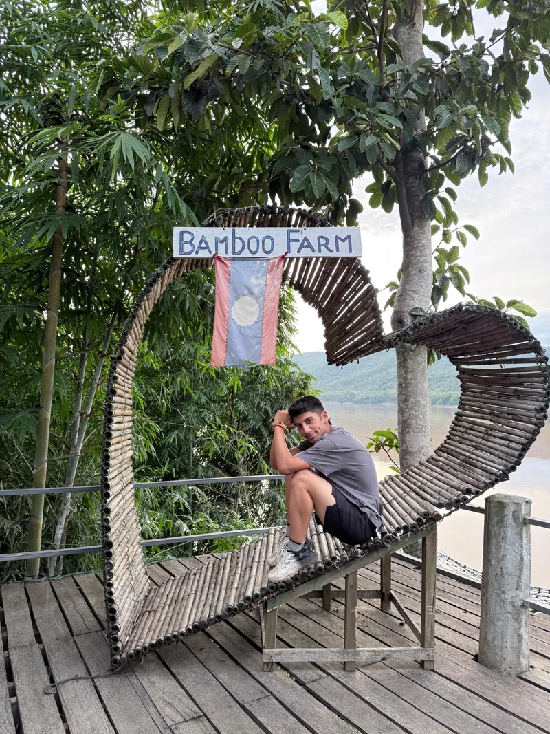 Yiğit นั่งบนที่นั่งไม้ไผ่โค้งใต้ป้าย Bamboo Farm และธงลาว มีแม่น้ำและเนินเขาด้านหลัง.