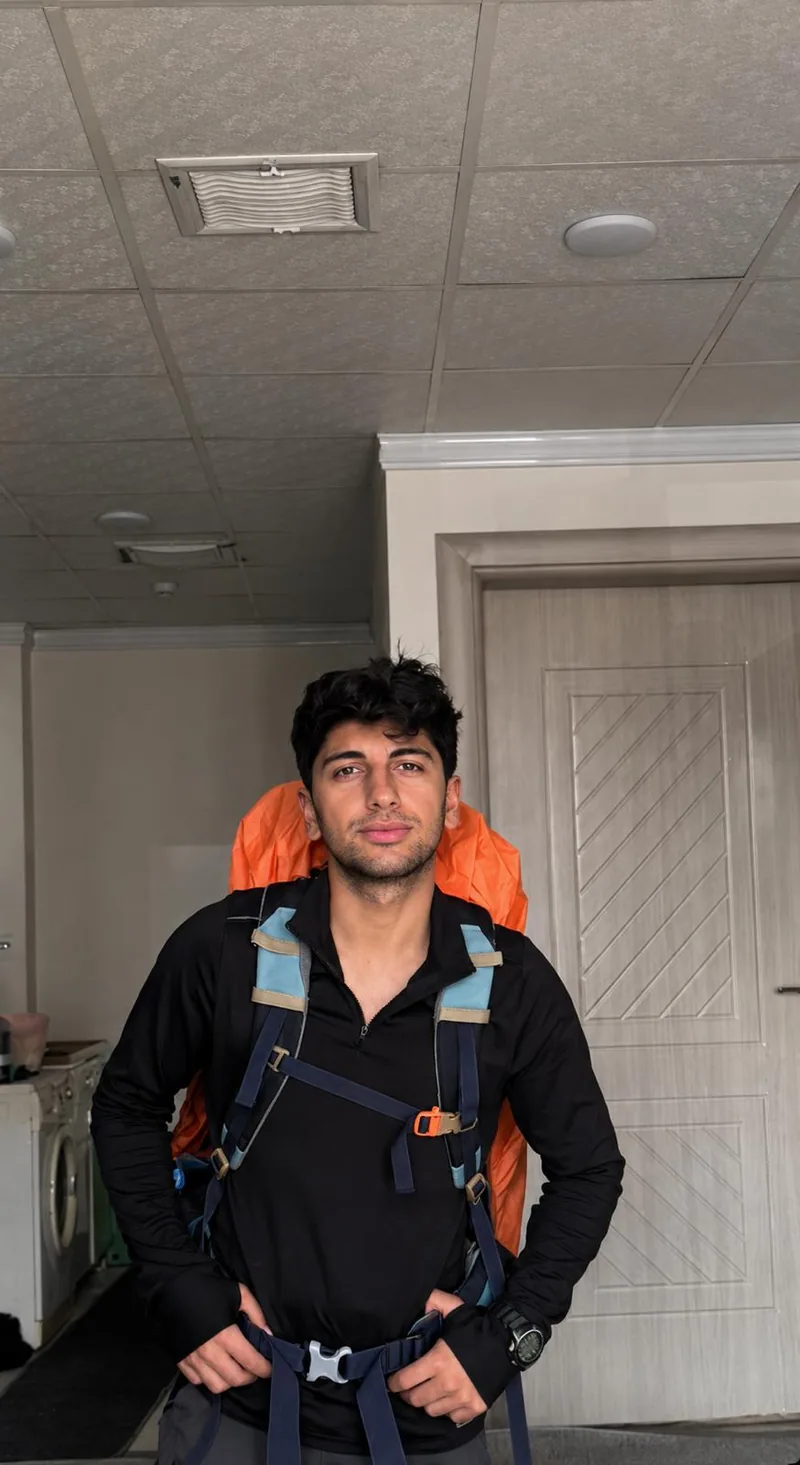 Yiğit ยืนอยู่ในอาคาร สวมเสื้อสีดำ และรัดสายหน้าอกของเป้เดินป่าที่มีผ้าคลุมกันฝนสีส้ม