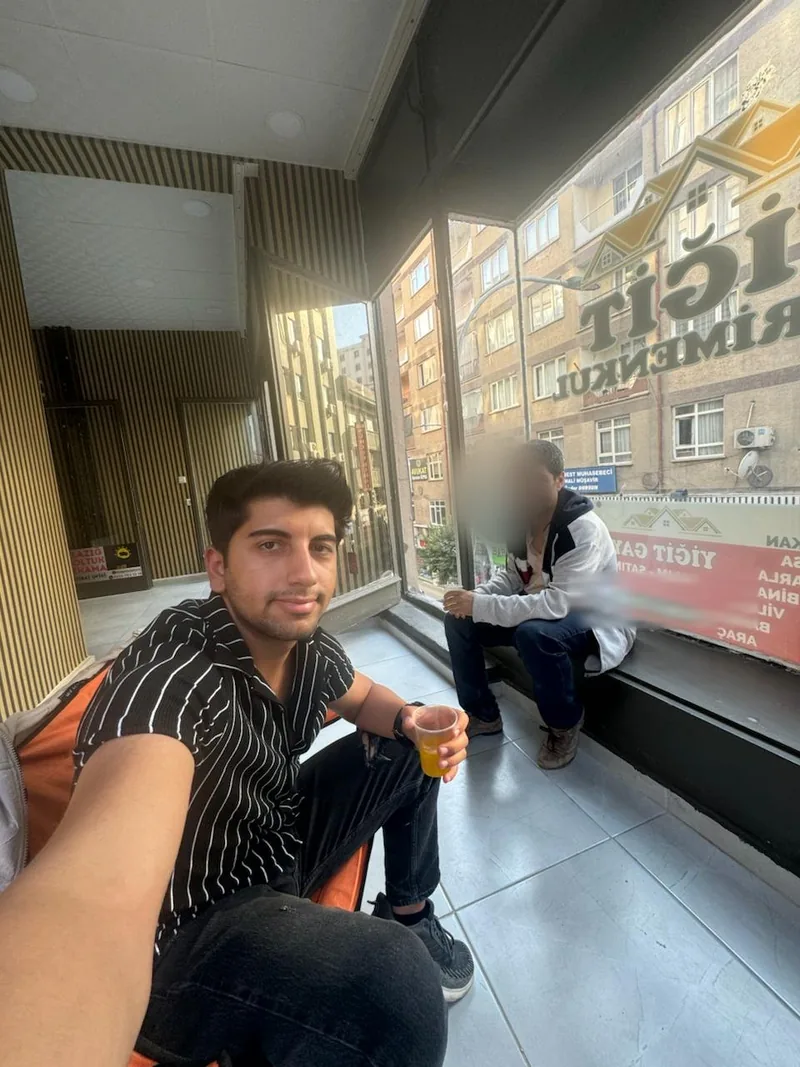 Yiğit ถ่ายเซลฟี่ในร่มข้างหน้าต่าง ขณะเพื่อนนั่งอยู่ใกล้ และมีตัวอักษรร้านด้านนอก