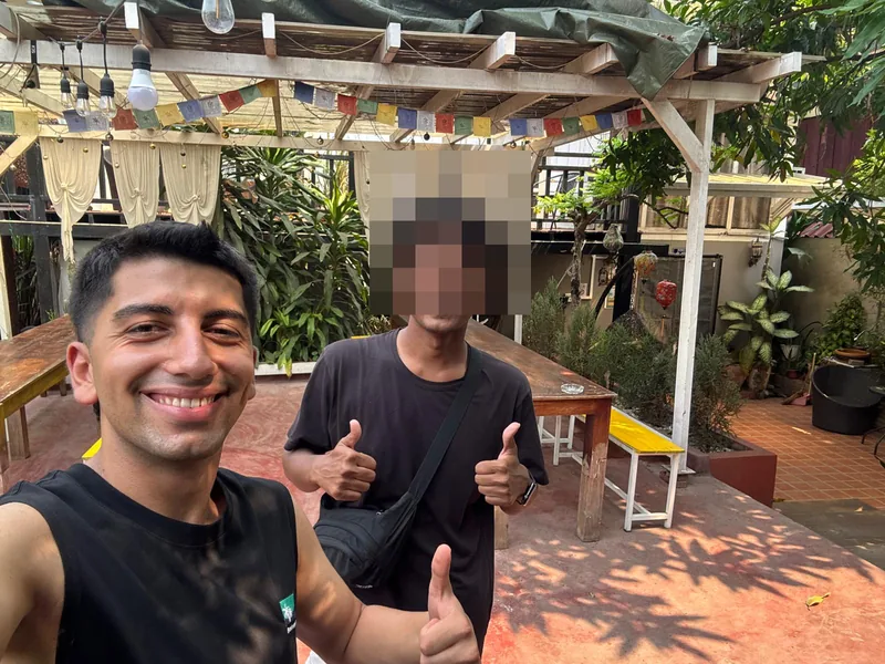 Yiğitและเพื่อนยิ้มและชูนิ้วโป้งในลานร่มรื่นใต้ธงมนต์และไฟประดับ.
