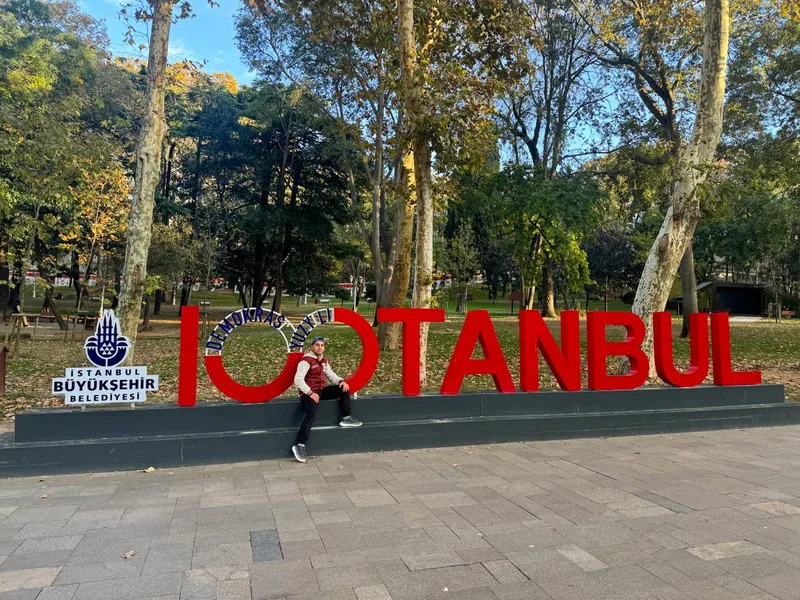 Yiğit นั่งในโค้งประติมากรรมตัวอักษร ISTANBUL สีแดงที่สวน Demokrasi Meydanı อิสตันบูล