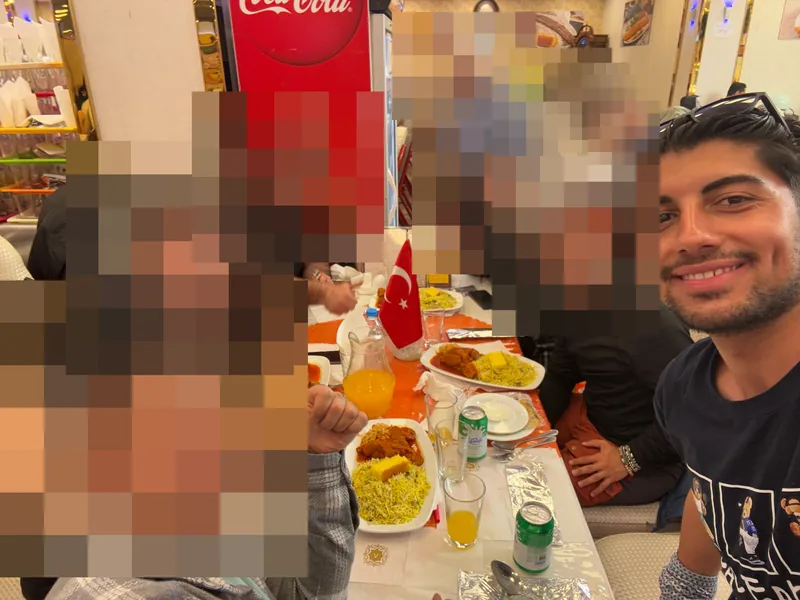 Yiğit ถ่ายเซลฟี่กับเพื่อนที่โต๊ะร้านอาหาร ข้างธงตุรกีและจานอาหาร.