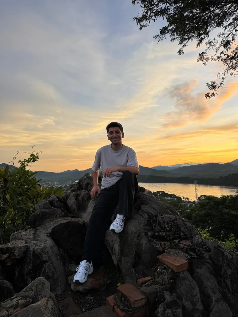 ยีกิตยิ้มขณะนั่งบนโขดหินเหนือแม่น้ำและภูเขาตอนพระอาทิตย์ตก