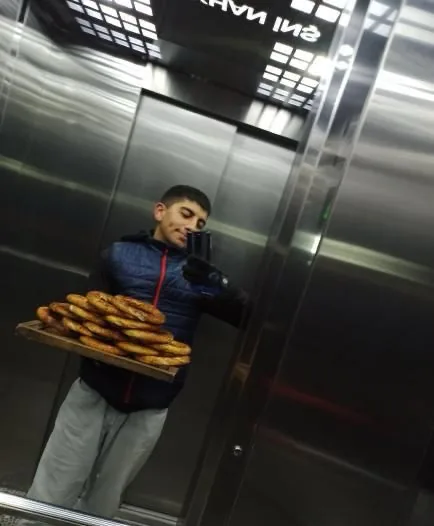 Yiğit holding a tray of simit, in an elevator mirror